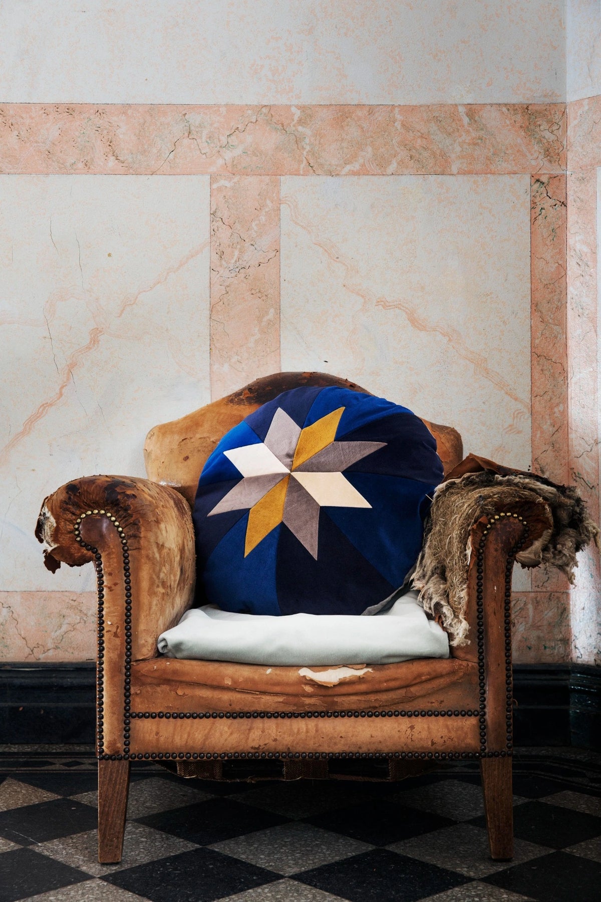 A round navy blue throw pillow with a geometric star pattern in yellow, beige, and white, styled on a brown leather armchair with animal fur accents against a marble wall.